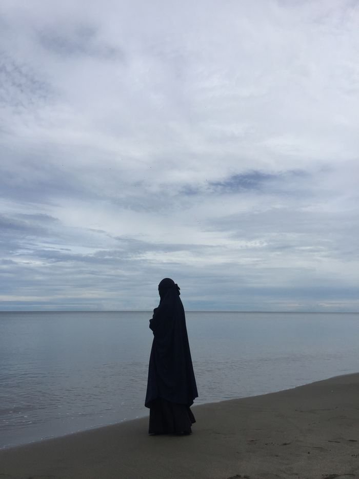 A woman in a hijab walks along a tranquil beach, reflecting against a vast seascape.